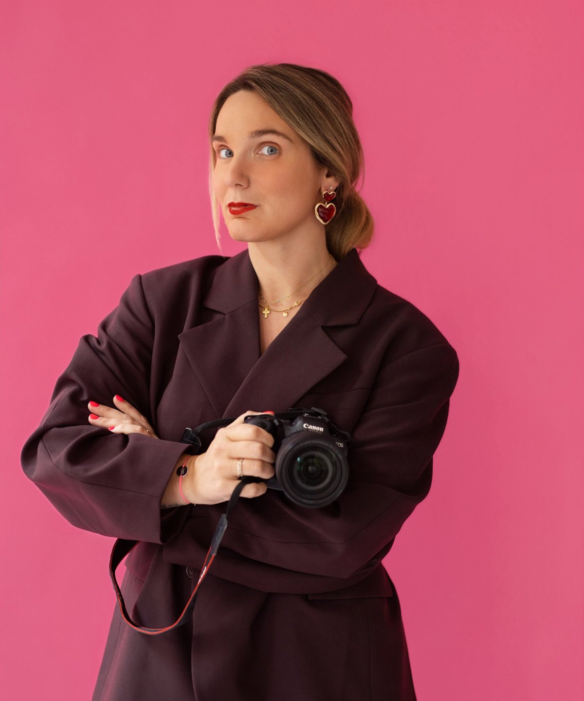 Marie Cotrel en blazer bordeaux sur fond rose, appareil photo Canon à la main