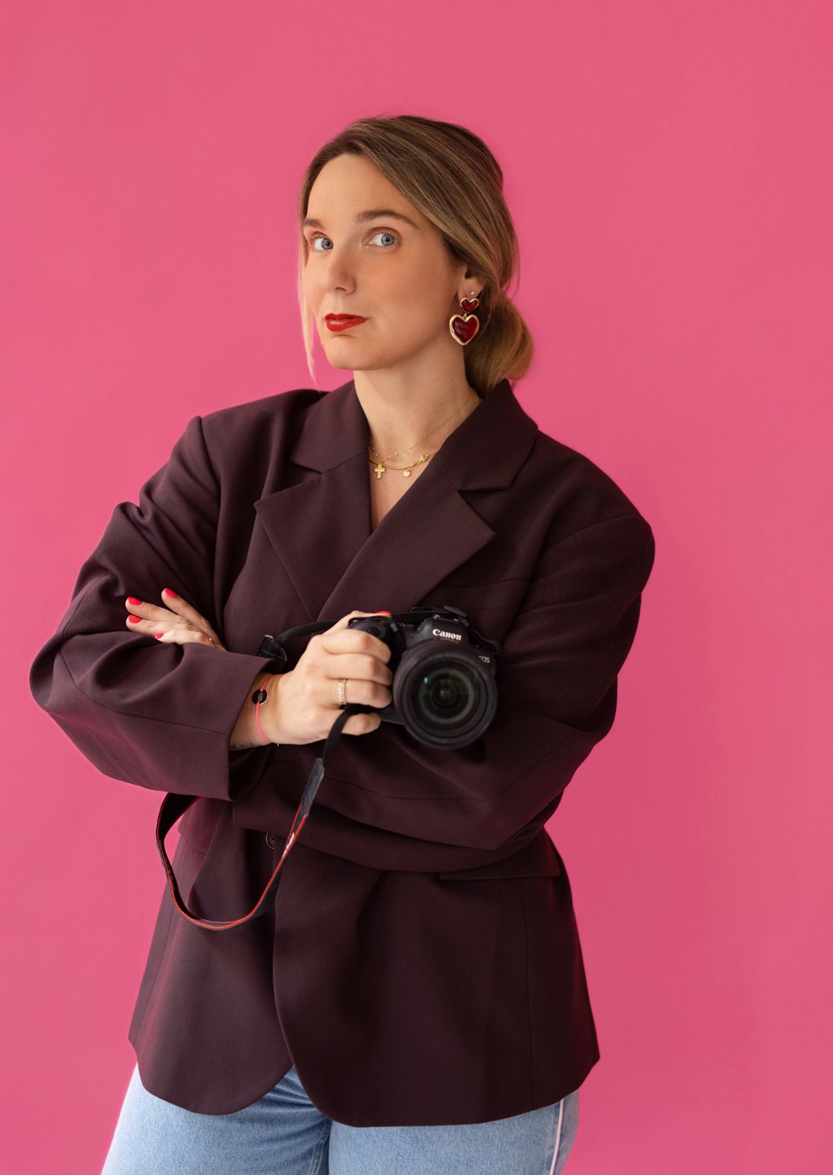 Marie Cotrel, photographe à Lyon, en blazer bordeaux sur fond rose, appareil photo Canon à la main